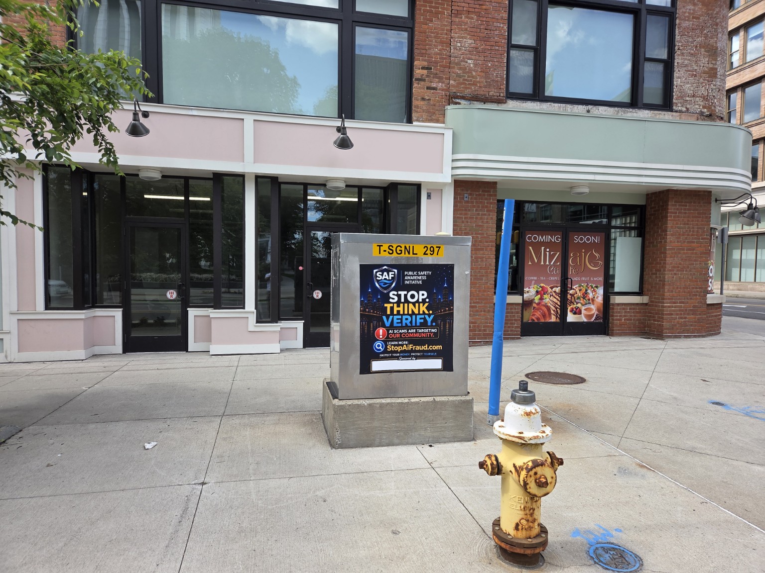 Close-up of the SAF Stop. Think. Verify. public safety sign on a traffic signal box at Main & Clinton in Downtown Rochester, showing the SAF shield logo, warning message, and StopAiFraud.com URL clearly visible to passersby.